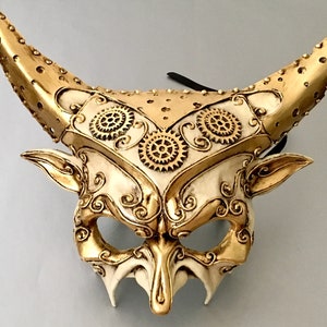 May include: A gold and white masquerade mask with horns and intricate details. The mask has a steampunk design with gears and swirls.