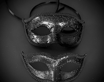 May include: Two black masquerade masks with intricate designs and glitter accents. The masks are made of plastic and have elastic straps.