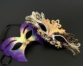 May include: Two masquerade masks, one gold and one purple, with intricate designs and rhinestones. The masks are connected by a black ribbon.
