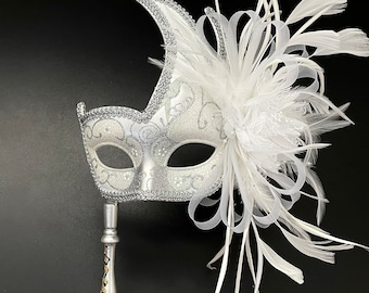 May include: A silver masquerade mask with white feathers and a stick handle. The mask has a delicate floral design and glitter accents.