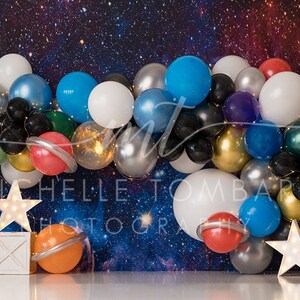 May include: A space-themed balloon arch with silver, gold, blue, red, black, and white balloons. The arch is decorated with small lights and planets. There are two white star-shaped lights on stands in front of the arch.