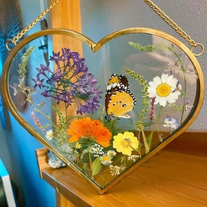 May include: Heart-shaped glass frame with a gold metal border, filled with dried flowers and a butterfly. The arrangement includes purple, orange, yellow, and white flowers. A gold chain is attached for hanging. The frame is approximately 8 inches wide.