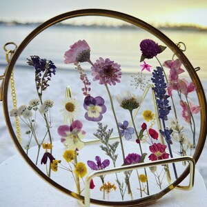 May include: A round, gold-toned framed glass display filled with an assortment of dried flowers. The flowers are in various colors, including purple, pink, yellow, and white. The frame has a small chain for hanging. The background is a blurred beach scene.
