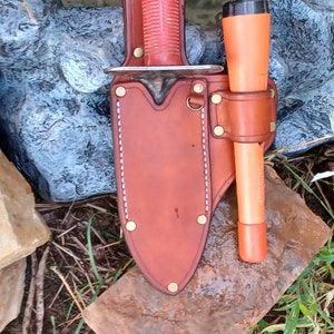 May include: A brown leather sheath with a red-handled knife and a flashlight. The sheath has white stitching and brass rivets. The knife and flashlight are both in the sheath.