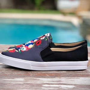 Puede incluir: Zapatillas sin cordones con un estampado floral azul marino en la parte superior, paneles laterales negros y suela blanca. El dise&ntilde;o floral incluye flores rojas, blancas y azules. Los zapatos est&aacute;n sobre una superficie de madera.