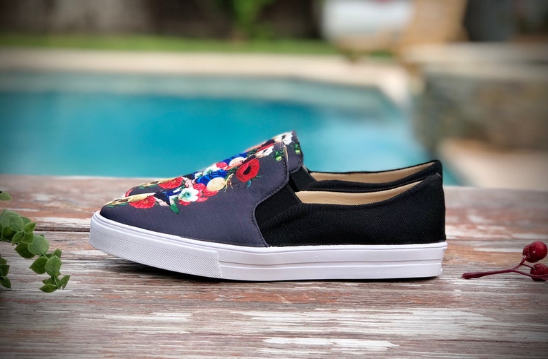 Puede incluir: Zapatillas sin cordones con un estampado floral azul marino en la parte superior, paneles laterales negros y suela blanca. El dise&ntilde;o floral incluye flores rojas, blancas y azules. Los zapatos est&aacute;n sobre una superficie de madera.