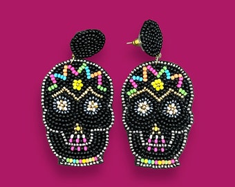 May include: A pair of black beaded skull earrings with colorful accents. The skulls have white eyes, pink noses, and a colorful design on the top of the head.