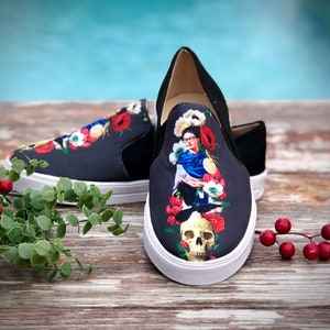 Puede incluir: Un par de zapatillas negras sin cordones con suela blanca. Las zapatillas presentan un dise&ntilde;o floral colorido y un retrato de una mujer con una calavera. Los zapatos est&aacute;n sobre una superficie de madera.