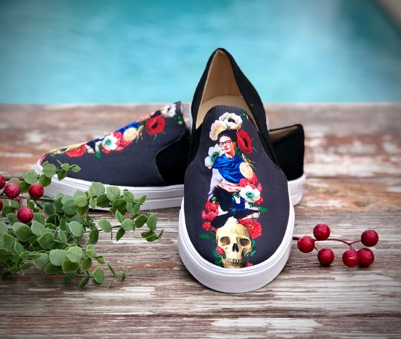 Puede incluir: Un par de zapatillas negras sin cordones con suela blanca. Las zapatillas presentan un dise&ntilde;o floral colorido y un retrato de una mujer con una calavera. Los zapatos est&aacute;n sobre una superficie de madera.