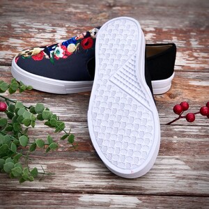 Puede incluir: Un par de zapatillas sin cordones. Un zapato presenta un dise&ntilde;o floral de calavera azul oscuro con suela blanca. El otro zapato es negro liso con suela blanca. Las suelas son blancas con un patr&oacute;n texturizado.