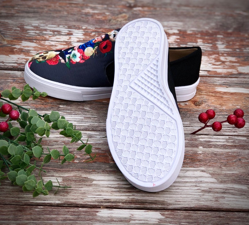 Puede incluir: Un par de zapatillas sin cordones. Un zapato presenta un dise&ntilde;o floral de calavera azul oscuro con suela blanca. El otro zapato es negro liso con suela blanca. Las suelas son blancas con un patr&oacute;n texturizado.