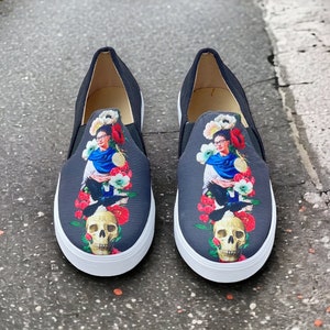 Puede incluir: Un par de zapatillas sin cordones negras con suela blanca. La parte superior de cada zapato presenta un gr&aacute;fico colorido de una mujer, flores, una calavera y p&aacute;jaros. Los zapatos tienen una textura similar a la lona.