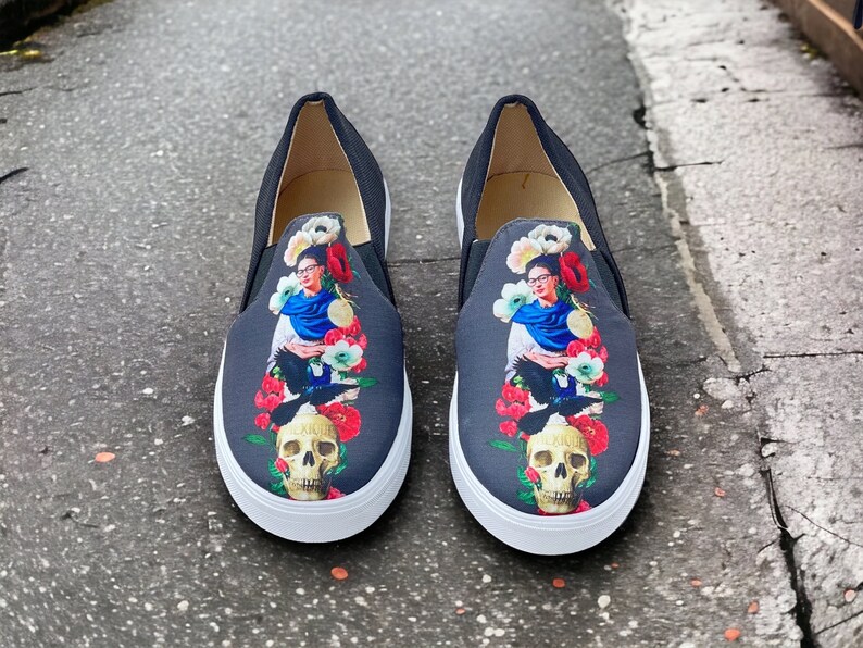 Puede incluir: Un par de zapatillas sin cordones negras con suela blanca. La parte superior de cada zapato presenta un gr&aacute;fico colorido de una mujer, flores, una calavera y p&aacute;jaros. Los zapatos tienen una textura similar a la lona.