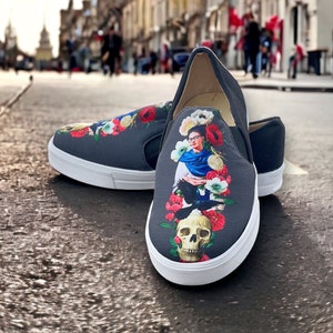 Puede incluir: Zapatillas grises oscuras con suelas blancas. Los zapatos presentan un dise&ntilde;o floral colorido con un retrato de mujer, una calavera y un p&aacute;jaro. Los zapatos est&aacute;n en una calle pavimentada.