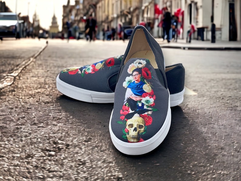 Puede incluir: Zapatillas grises oscuras con suelas blancas. Los zapatos presentan un dise&ntilde;o floral colorido con un retrato de mujer, una calavera y un p&aacute;jaro. Los zapatos est&aacute;n en una calle pavimentada.