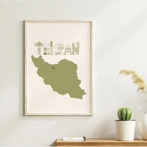 May include: A framed art print featuring a map of Iran in olive green with the city name "Tehran" above. The artwork is displayed on a white wall, with a wooden shelf below holding plants and decorative items.