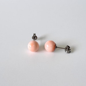 Puede incluir: Un par de pendientes de botón con cuentas redondas y lisas de color rosa pálido. Cada cuenta está montada sobre un poste de metal oscuro con un cierre de mariposa a juego, creando un diseño sencillo y limpio para estas piezas.