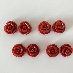Puede incluir: Seis objetos rojos tallados en forma de rosa están dispuestos sobre una superficie blanca. Las rosas tienen pétalos detallados y un acabado brillante. Los objetos parecen ser componentes de joyería o elementos decorativos.