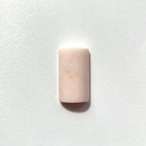 Puede incluir: Una piedra rectangular de color rosa pálido con sutiles variaciones de color. La piedra es lisa y pulida, con bordes redondeados. El objeto está sobre un fondo blanco, creando una estética limpia y minimalista.