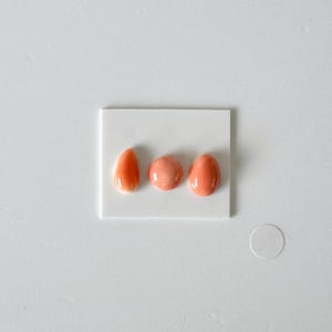 Op de afbeelding: Drie gladde, gepolijste, perzikkleurige cabochons horizontaal gerangschikt op een kleine witte rechthoekige kaart. De cabochons bestaan uit een druppelvorm, een cirkelvorm en een ovale vorm, allemaal met een glanzend, reflecterend oppervlak.