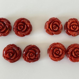 Puede incluir: Ocho cabujones de rosas rojas talladas. Los cabujones están dispuestos en dos filas de cuatro, cada uno con formaciones detalladas de pétalos. Las rosas son de un color rojo intenso y rico, y parecen estar hechas de un material pulido.