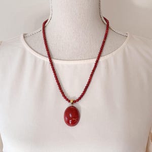 May include: A red beaded necklace with a large, oval pendant. The pendant and beads are a deep red color. The necklace is displayed on a white mannequin.