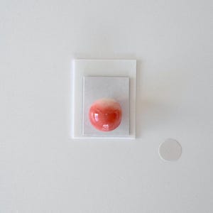 Può includere: Un oggetto sferico lucido con un gradiente dal rosso al bianco, appoggiato su una base rettangolare bianca. La base è stratificata su un rettangolo bianco più grande. Un piccolo oggetto circolare bianco è a destra.