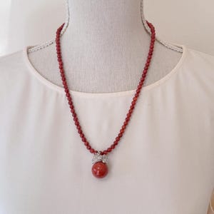 May include: A red coral bead necklace with a matching pendant. The pendant features a large, round coral bead held by a silver-colored metal setting with leaf-like details. The necklace is displayed on a white mannequin.