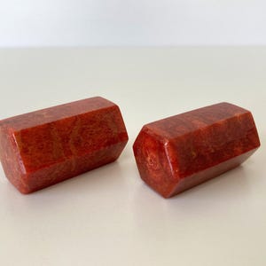 May include: Two red-orange, hexagonal-shaped objects with a glossy finish. The objects have a marbled appearance with darker red veining. They are displayed on a white surface.