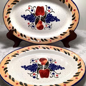 May include: Two white oval ceramic serving platters with a speckled glaze. The platters feature a hand-painted design of red apples, blue grapes, and green leaves on a white background. The platters have a brown, orange, and blue border.