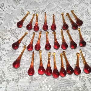 May include: A collection of twenty-three teardrop-shaped glass pendants. Each piece features a deep red, rounded base that transitions into a slender, translucent amber-colored neck, topped with a small circular loop for hanging or attachment.