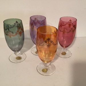 May include: Set of four glass drinking glasses with gold trim and a decorative design. The glasses are in shades of green, purple, pink, and orange.