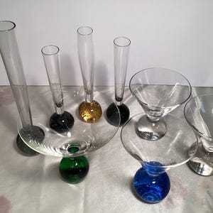 May include: A set of three ceramic mugs with a speckled, cream-colored glaze. Each mug features a different hand-painted design: a green leaf, a blue flower, and a yellow sun. The mugs have a simple, modern design.