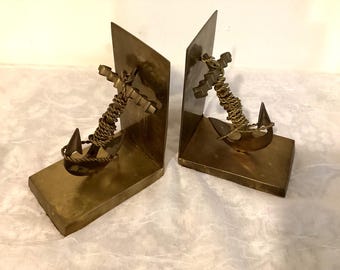 May include: A pair of brass bookends shaped like anchors with rope details. The bookends are on a white surface.