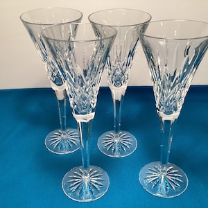 May include: Four clear crystal champagne flutes. Each flute has a tall, slender stem and a starburst design on the base. The bowls are etched with a diamond pattern. The flutes are arranged on a blue surface.