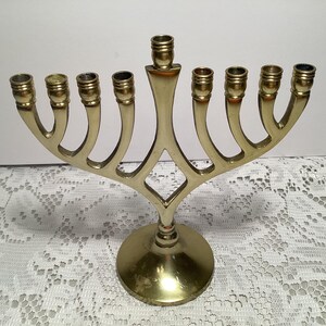 May include: A polished brass menorah featuring a central stem and eight curved arms extending outward, each topped with a cylindrical candle cup. The structure is supported by a circular base, showcasing a smooth, reflective metallic finish.