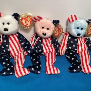May include: Three plush teddy bears with patriotic designs. Each bear has a different colored head: white, pink, and light blue. The bodies are split with red and white stripes on one side and navy blue with white stars on the other.