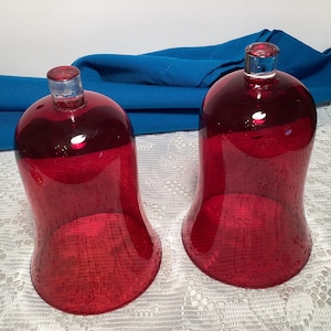 May include: Two ruby red glass candle holders with clear glass stoppers. The bell-shaped holders have a textured surface and are designed to hold candles. The background includes a blue cloth and a white lace tablecloth.
