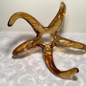 May include: A clear amber glass sculpture shaped like a starfish with five points. The sculpture has a swirl pattern throughout.