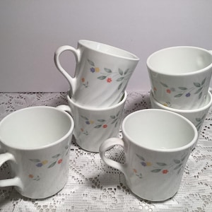May include: A set of five white ceramic mugs featuring a delicate pattern of small orange and yellow flowers with grey leaves. Each mug has a subtle spiral texture on the exterior surface and a curved handle, arranged in a stacked and grouped formation.