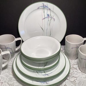 May include: A dinnerware set featuring white ceramic plates, bowls, and mugs with a delicate purple and green iris floral pattern and a thin green rim. The collection includes a large dinner plate, stacked smaller plates, a bowl, and four mugs.