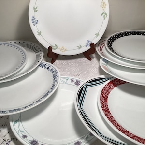 May include: A collection of white Corelle plates with various decorative borders. Designs include floral patterns, polka dots, and geometric shapes in colors like blue, black, and red. Some plates are stacked, others are displayed on a wooden stand.