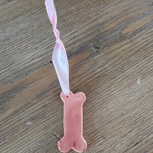 Könnte beinhalten: Ein rosa knochenförmiger Keramikschmuck mit einem Loch oben, befestigt an einem rosa Band. Der Schmuck hat einen hellrosa Farbton und eine glänzende Oberfläche. Der Hintergrund ist eine Holzoberfläche.