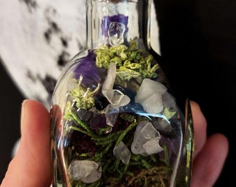 May include: A small clear glass bottle with a cork stopper containing a collection of green moss, dried purple flower petals, and various small tumbled clear and dark blue crystals layered inside the vessel to create a decorative display.