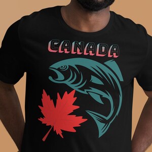 Op de afbeelding: Zwart T-shirt met een turquoise visafbeelding, een rode esdoornblad en het woord "CANADA" in een roze en witte gradiënt. Het T-shirt heeft een ronde hals.