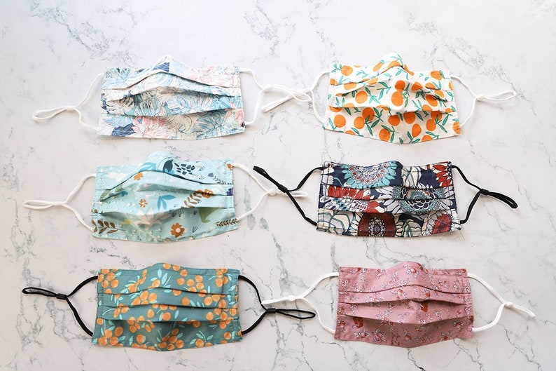 May include: Six different fabric face masks with various patterns. The masks are pleated and have white elastic ear loops. The patterns include floral, orange slices, and abstract designs.