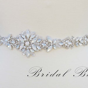 May include: A white bridal belt with a silver and white crystal design. The belt is adorned with a large cluster of crystals in the center.