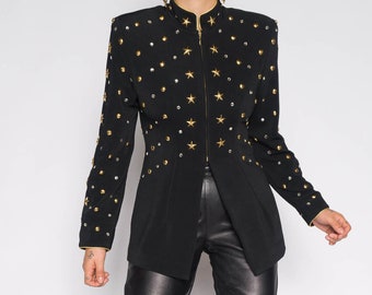 Op de afbeelding: Een zwart blazer met gouden ster-versieringen en een ritssluiting. De blazer heeft een staande kraag en lange mouwen. Het model draagt een zwarte leren broek.