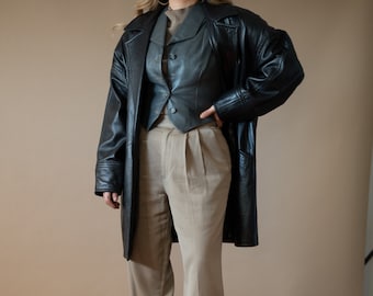 Könnte beinhalten: Eine Frau trägt eine schwarze Lederjacke, eine schwarze Lederweste und eine beige Hose. Sie steht auf einem Holzboden.