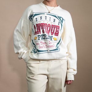 May include: A white sweatshirt with a vintage-style graphic print that says "Genuine Antique Persian" with a red and blue design. The sweatshirt is paired with cream-colored wide-leg pants.